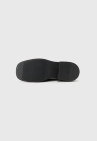 Semelle de chaussure en caoutchouc noir avec une surface texturée, dotée de rainures horizontales et verticales, de forme effilée et sans marquage visible.