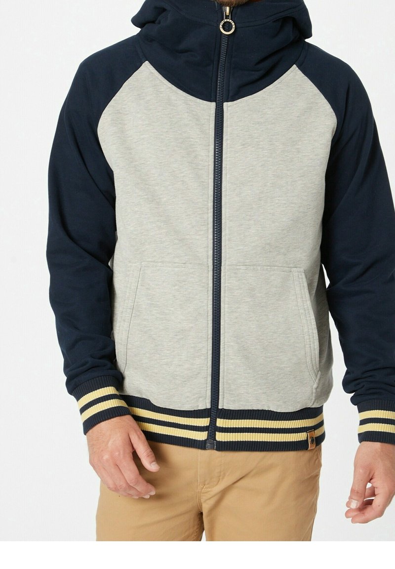Homme portant un sweat à capuche zippé avec manches et capuche bleu marine, torse gris clair, et poignets et ourlet rayés bleu marine et jaune, assorti à un pantalon beige.