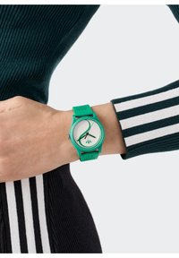 Roheline silikoonkell ümmarguse valge näoga, minimalistlike tunni ja minuti kätega ning logo allservas; kantud randmel triibulisest sviiterist.