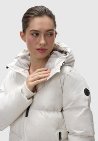 Giacca a vento bianca con cappuccio, dotata di una morbida fodera interna grigia, polsini a coste e un logo circolare nero sulla manica.