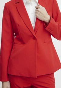 Blazer rouge à coupe ajustée, fermeture à un bouton et deux poches avant. Porté sur une chemise blanche à col. Texture lisse.