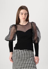 Zwarte top met doorschijnende, poffende mouwen versierd met witte stippen; strakke lijfje structuur. Gepaard met een zwart-wit houndstooth rok.