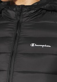 Chaqueta negra acolchada con cremallera frontal y logo blanco de "Champion" en el pecho izquierdo, llevada por una persona.