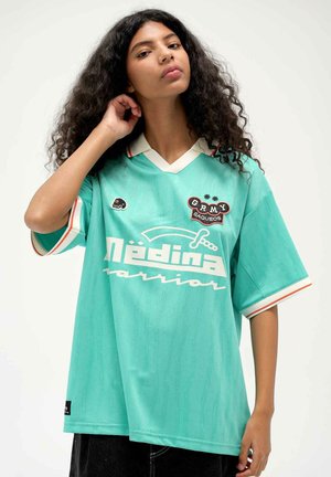 Junge Person mit lockigem Haar, die ein lockeres tealfarbenes Medina Warrior-Shirt mit weißem Kragen und Aufnähern trägt und mit einer Hand nahe am Ohr posiert.