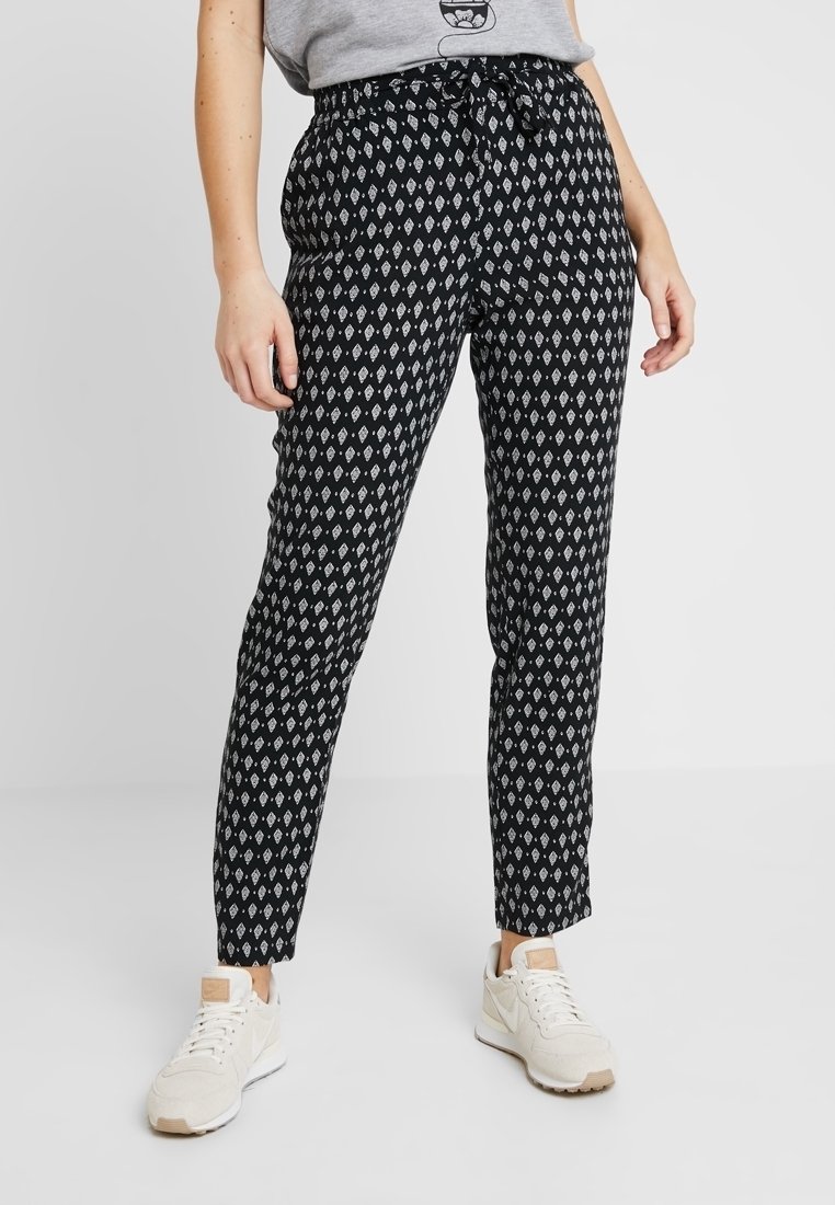 Femme portant un pantalon noir avec des motifs en losanges blancs, un t-shirt gris rentré et des baskets blanches, debout devant un fond uni.