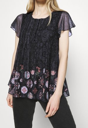 Femme portant un chemisier noir transparent à manches courtes avec des boutons sur le devant et des motifs floraux circulaires à l'ourlet, associé à un pantalon foncé.