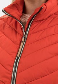 Czerwona pikowana kurtka z wysokim kołnierzem i zamkiem błyskawicznym z przodu. Posiada beżową lamówkę wzdłuż zamka i kontrastową teksturowaną podszewkę wewnętrzną.