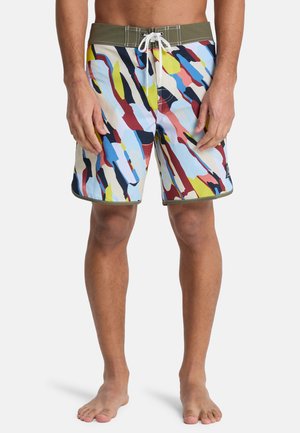 Mann trägt knielange Badehose mit mehrfarbigem abstraktem Muster, olivgrünem Bund und weißem Kordelzug, steht barfuß.