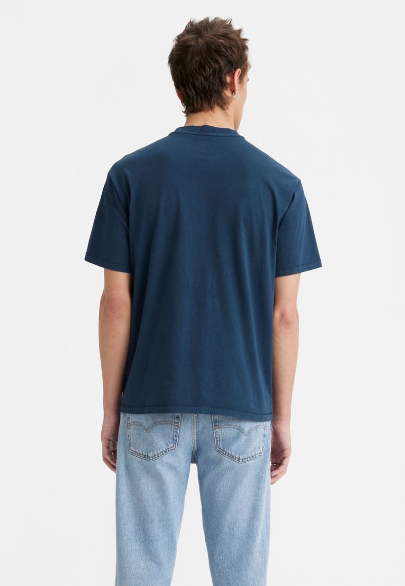 Homme debout, de dos, portant un t-shirt uni bleu foncé à manches courtes et un jean bleu clair, sur un fond blanc uni.