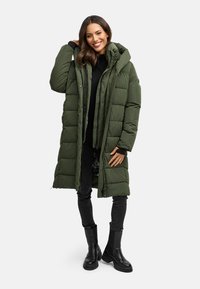 Lange, olijfgroene pufferjas met een capuchon, rits- en drukknoopsluitingen, een gestikt ontwerp en gecombineerd met zwarte broek en forse zwarte laarzen.