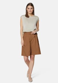 Lichtbeige mouwloze top gecombineerd met bruine geplooide shorts, voorzien van een riem. Model draagt naakte hakken en staat tegen een witte achtergrond.