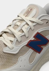 Gros plan sur une basket beige et blanche en tissu mesh, lacets crème, logo "N" bleu avec bordure rouge, et languette rembourrée avec texte étiquette.