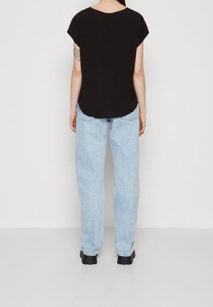 Persoon staand met de rug naar de camera, draagt een loszittende zwarte top met korte mouwen, lichtblauwe spijkerbroek, zwarte schoenen en een tatoeage van een spinnenweb op de linkerarm.