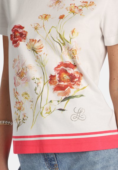 White short-sleeve shirt with large red and pink floral design, green stems, and coral pink stripes at the hem, worn with blue jeans.