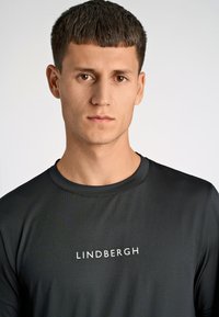 Svart fukttransporterande träningsskjorta med rund halsringning, med en texturerad yta och "LINDBERGH"-logotypen i vitt över bröstet.