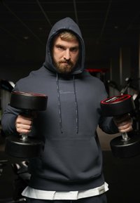 Grijze hoodie met een voorzak, voorzien van een gevoerde capuchon. De persoon houdt twee zwarte dumbbells met rode accenten vast, wat een sportschoolomgeving benadrukt.