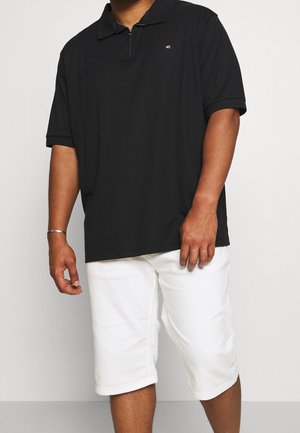 Mężczyzna ubrany w czarną koszulkę polo z krótkim rękawem i białe szorty do kolan, stojący na jednolitym tle.