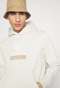 Felpa leggera beige con cappuccio, dotata di tasca frontale e logo bianco con "Calvin Klein Jeans". Accompagnata da un cappello a secchiello color sabbia. Texture liscia.