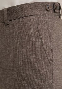 Bruine textuurbroek met een strakke afwerking, voorzien van twee knopen bij de tailleband en zijzakken voor functionaliteit.