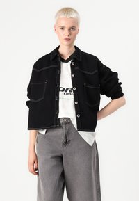 Casaco oversized preto com costuras em contraste, destacando dois bolsos no peito. Usado sobre uma t-shirt branca e calças largas cinzas.