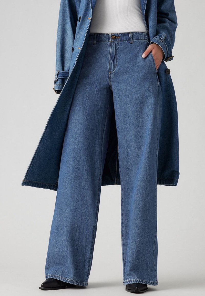 Personne portant un jean bleu à jambes larges, un haut blanc et un long manteau en jean bleu, avec une main dans la poche et des chaussures noires visibles.