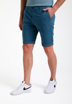 Teal katoenen shorts met opgerolde mouwen, voorzien van zijzakken en een knoopsluiting. Gecombineerd met witte sneakers met zwarte accenten.