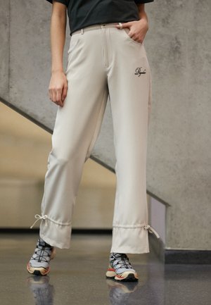Μπεζ παντελόνια σε ίσια γραμμή από απαλό ύφασμα, με πλαϊνές τσέπες και κορδόνια στο τελείωμα, συνδυασμένα με αθλητικά παπούτσια με σχέδιο.
