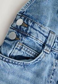 Gros plan sur la poche d'un jean en denim bleu clair avec deux rivets argentés et des détails de couture visibles.