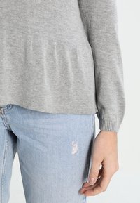 Grauer Strickpullover mit Peplum-Design und gerippten Bündchen, kombiniert mit hellblauen, verwaschenen Jeans, die ein kleines Loch am Oberschenkel haben.