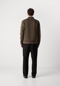 Olive green bomber jacket with a ribbed hem, collar, and cuffs; worn with black trousers and features a logo patch on the left sleeve.