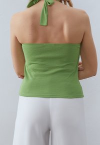 Žena v zeleném halter topu svázaném za krkem a bílých kalhotách s vysokým pasem, zobrazená zezadu na jednoduchém světlém pozadí.