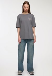 Szara bawełniana koszulka z okrągłym dekoltem i krótkimi rękawami, połączona z jasnoniebieskimi szerokimi spodniami dżinsowymi. Koszulka ma małe nadrukowane logo.