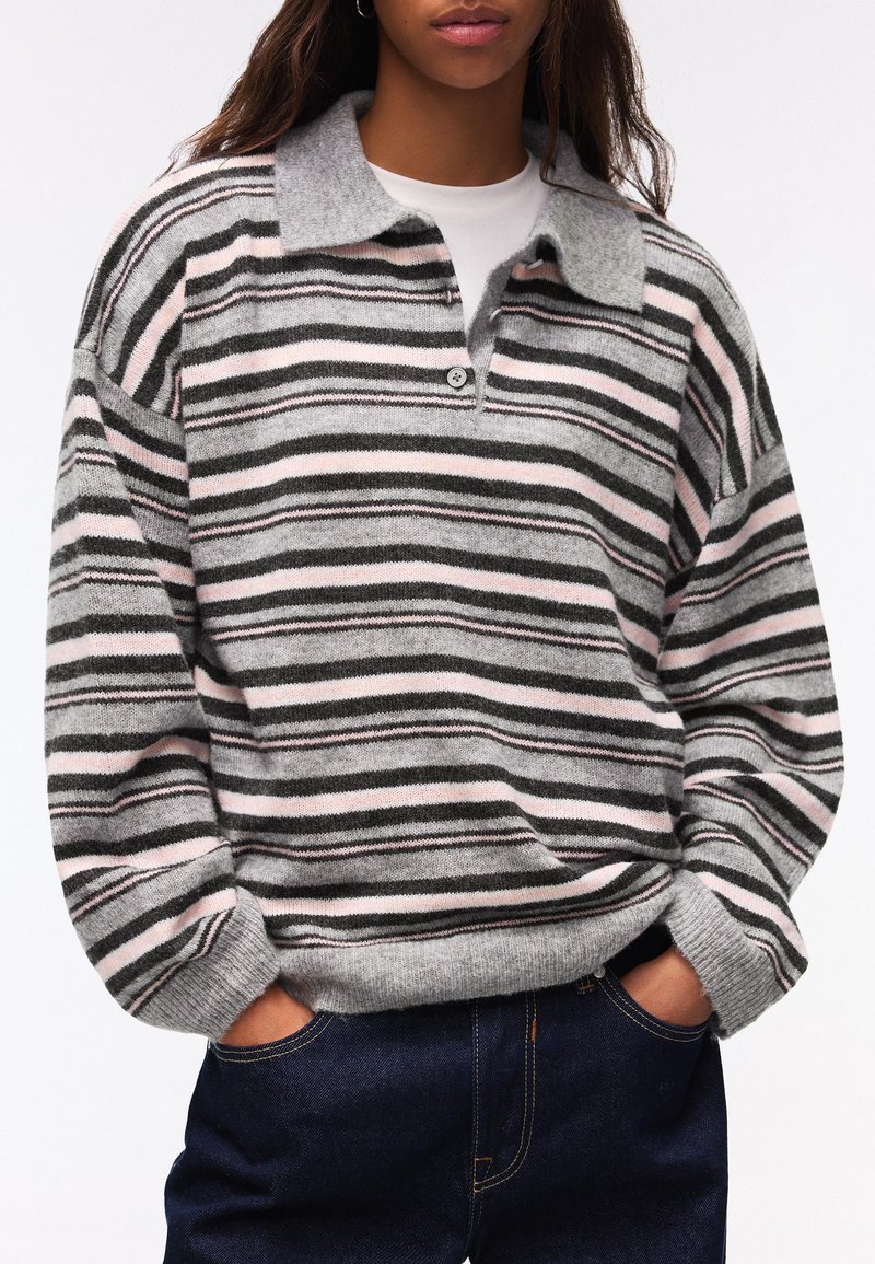 Persona che indossa un maglione a righe grigio, rosa e nero con colletto e camicia bianca interna, con le mani nelle tasche di jeans blu scuro.