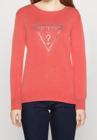 Maglione color corallo realizzato in tessuto morbido con maniche lunghe, scollo rotondo e un dettaglio triangolare con logo argentato sul petto.