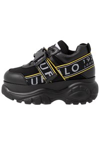 Tjock svart sneaker med nät- och syntetöverdel, med gula och vita ränder samt ett framträdande spänne. Tjock gummisula.