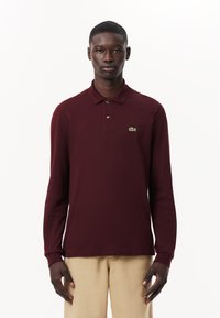 Burgundy långärmad polo med krage, med tre knappar och en grön broderad logotyp på vänstra bröstområdet.
