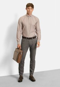 Hellbraunes Hemd mit Knöpfen, graue gemusterte Hose, dunkler Gürtel, braune Aktentasche und braune Schuhe. Das Modell steht vor einem einfarbigen Hintergrund.