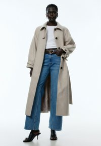 Lang beige wollen jas met grote zwarte knopen, gecombineerd met hoge blauwe jeans. De outfit wordt compleet gemaakt met zwarte haksandalen.