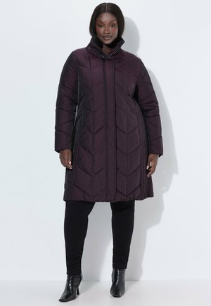 Mujer de pie con un abrigo acolchado oscuro de longitud hasta la rodilla, pantalones negros ajustados y botines negros de tacón contra un fondo blanco liso.