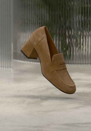 Bruine suède loafers met een vierkante neus en een kleine blokhak, voorzien van een decoratieve riem aan de voorkant. Soepele textuur, minimalistisch ontwerp.