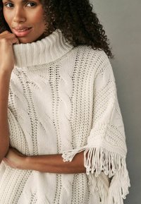Cremefarbenes gestricktes Poncho mit hohem Rollkragen, ausgestattet mit Zopfmuster und Fransenakzenten an den Ärmeln.