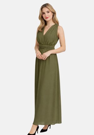 Femme portant une robe maxi sans manches vert olive avec taille froncée et des talons noirs, debout les mains jointes.