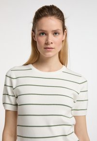 Witte gebreide top met groene horizontale strepen, ronde hals en korte mouwen. Zachte textuur en aansluitend ontwerp. Geen opvallende accessoires of details.