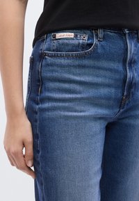 Μπλε τζιν από τζιν υλικό με ψηλή μέση, πέντε τσέπες και κόκκινη και λευκή ετικέτα Calvin Klein στο μπροστινό μέρος. Λείο υλικό με λεπτομέρειες ξεθωριάσματος.