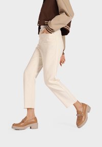 Scarpe in pelle marrone con suola spessa, dotate di una fascetta decorativa e una fibbia. Indossate con pantaloni color crema e una giacca marrone.