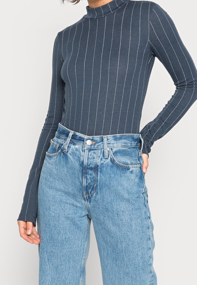 Femme portant un col roulé gris foncé à manches longues avec des rayures verticales blanches et un jean bleu clair taille haute.