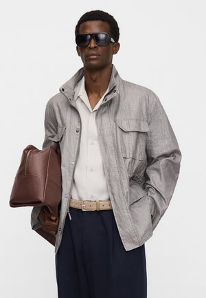 Man met een grijze jas, wit overhemd, donkerblauwe broek, beige riem, grote zwarte zonnebril, en een bruine leren tas onder de arm.