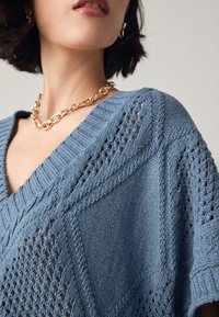 Pulover tricotat de culoare albastru deschis, cu decolteu în V, modele texturate și croială lejeră. Accesorizat cu un colier gros din lanț auriu.