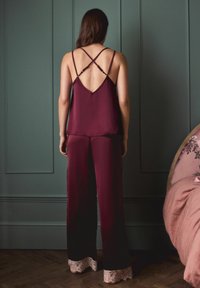Burgunderfarbene Satin-Camisole mit gekreuzten Trägern, kombiniert mit weiten Hosen mit Spitzenbesatz am Saum. Glatte, fließende Textur.