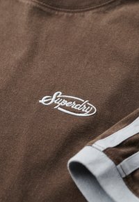 Camiseta de algodón marrón con un logo bordado en blanco de "Superdry", con rayas de acento gris en las mangas. Tejido suave y texturizado.